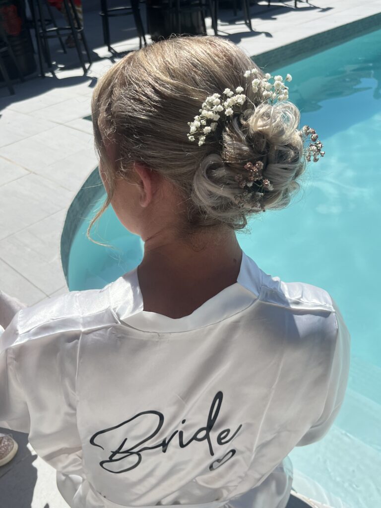 Vue de dos d’une mariée avec un chignon sophistiqué orné de petites fleurs blanches, coiffure réalisée par Les Coiffures de Mel, votre coiffeuse à domicile à La Destrousse.